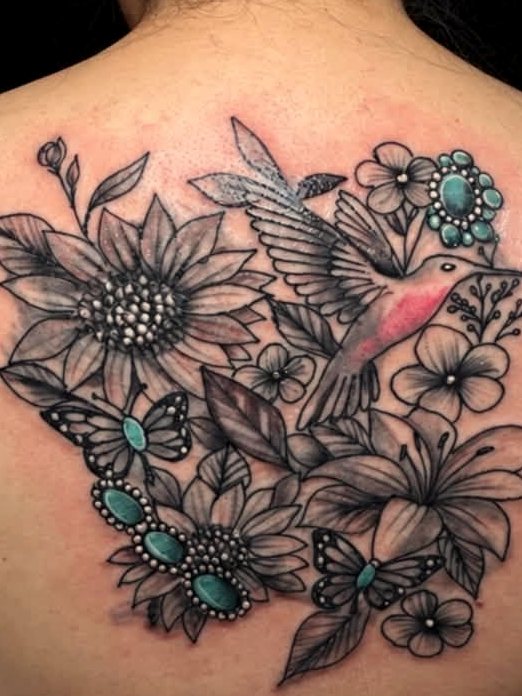 Black-and-grey back piece — a hummingbird among sunflowers and lilies, picked out with turquoise stones.